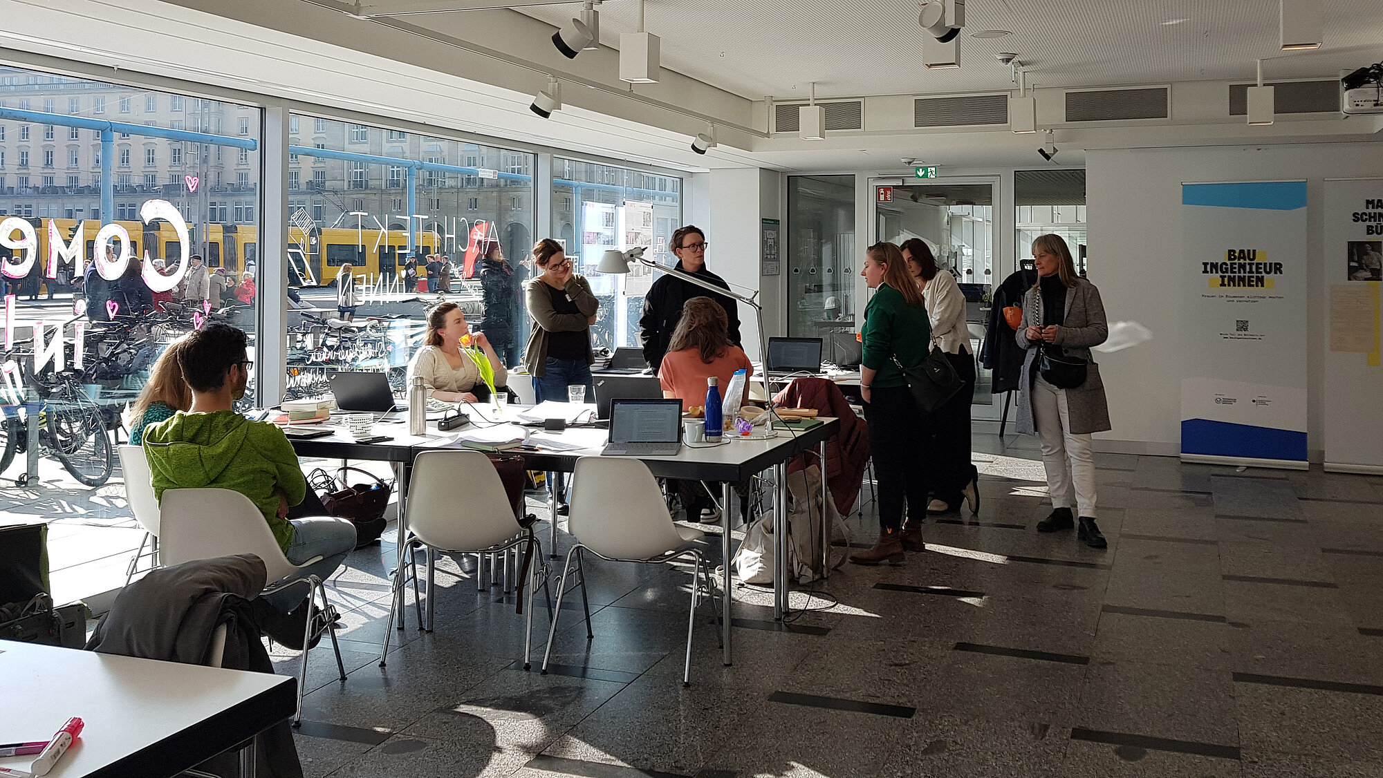 (Architektenkammer Sachsen) eine Gruppe von Frauen, sitzend und stehend, um Schreibtische und Stühle versammelt und in verbalem Austausch. Die linke Seite des Raumes ist von raumgroßen Glasscheiben flankiert, die das Stadtleben draußen zeigen.